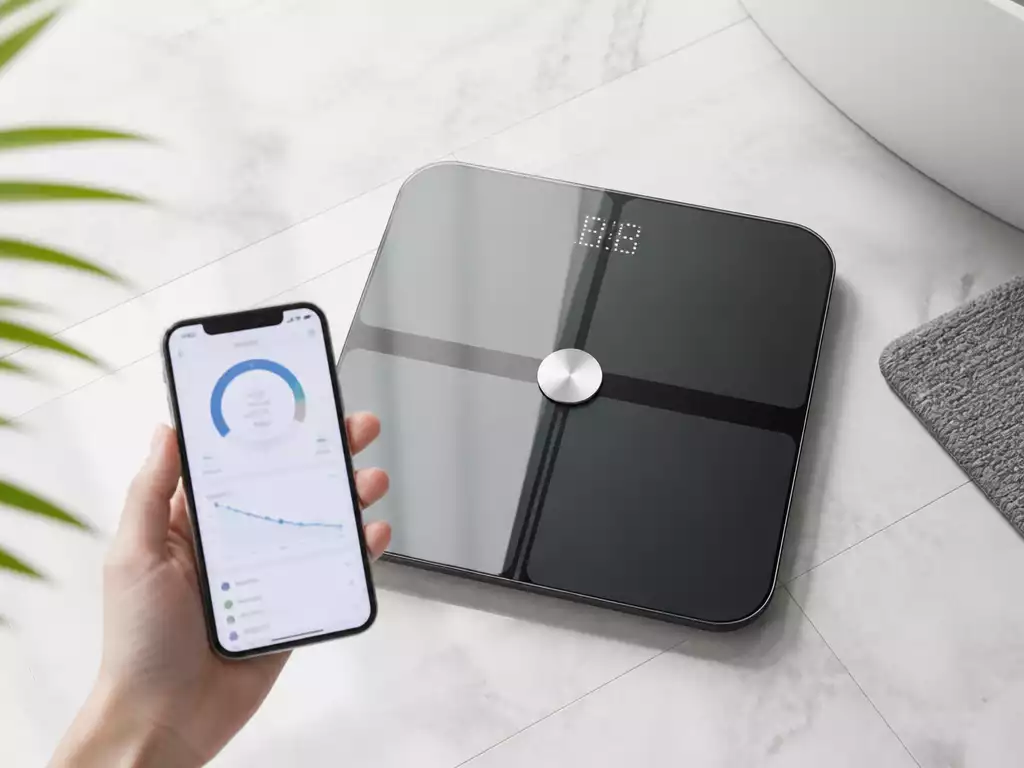 Modern iHome smart scale on a bathroom floor, displaying weight, with a smartphone showing a health app connected via Bluetooth