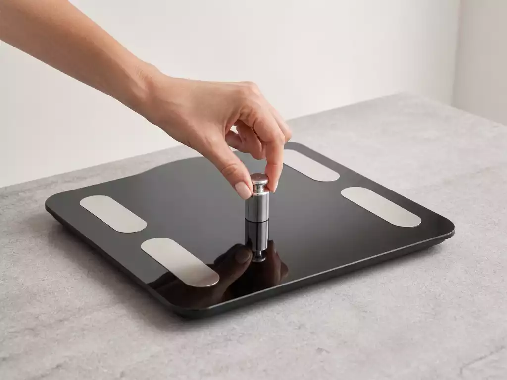Modern Beurer scale on a flat, stable surface with a hand placing a calibration weight on it