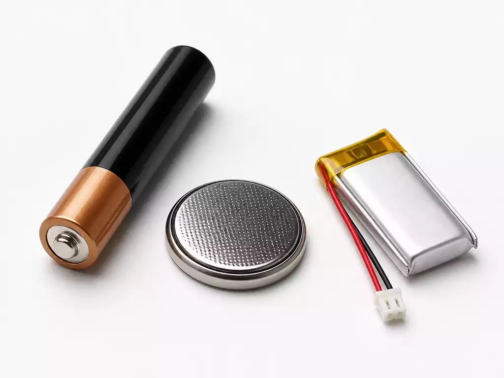 Close-up of different types of batteries (AAA, CR2032, and a small rechargeable lithium battery) arranged neatly on a clean white surface with soft studio lighting