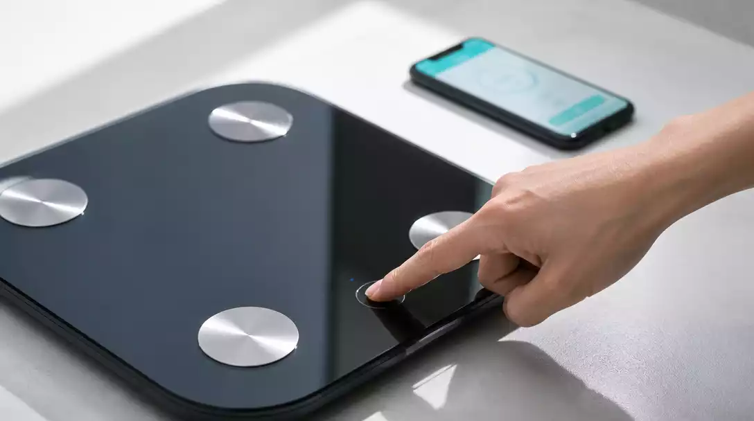 Person's hand pressing a reset button on a smart scale, with a smartphone displaying a health app in the background