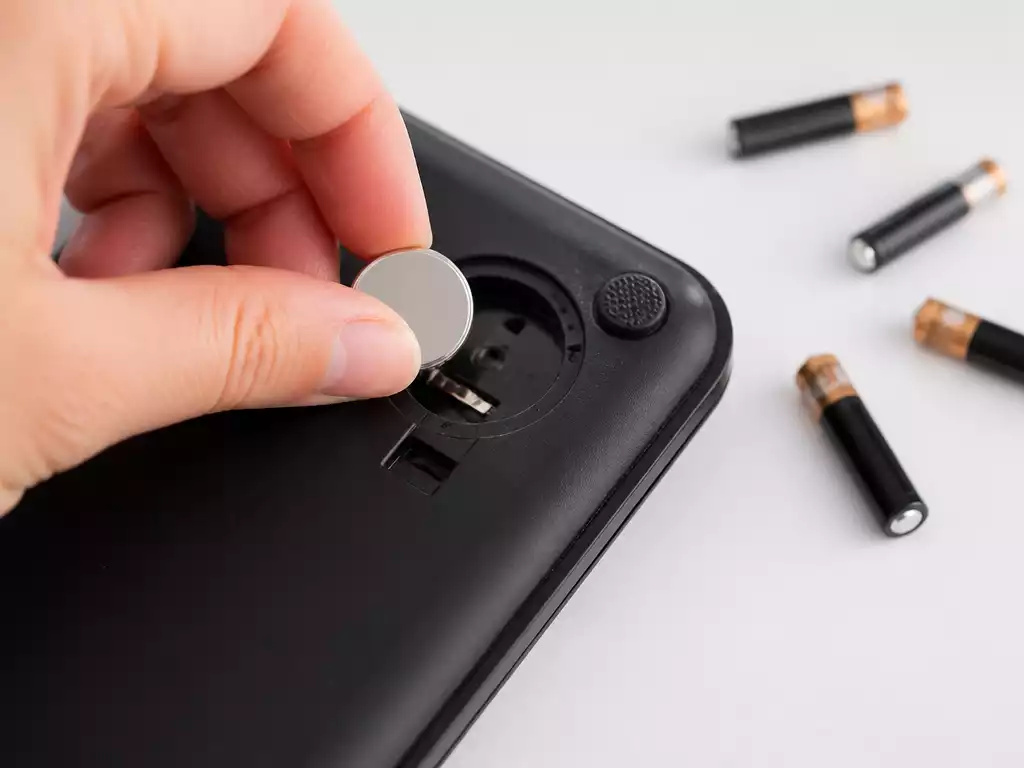 Close-up of a hand placing a CR2032 battery into a small Etekcity scale, with AAA batteries in the background