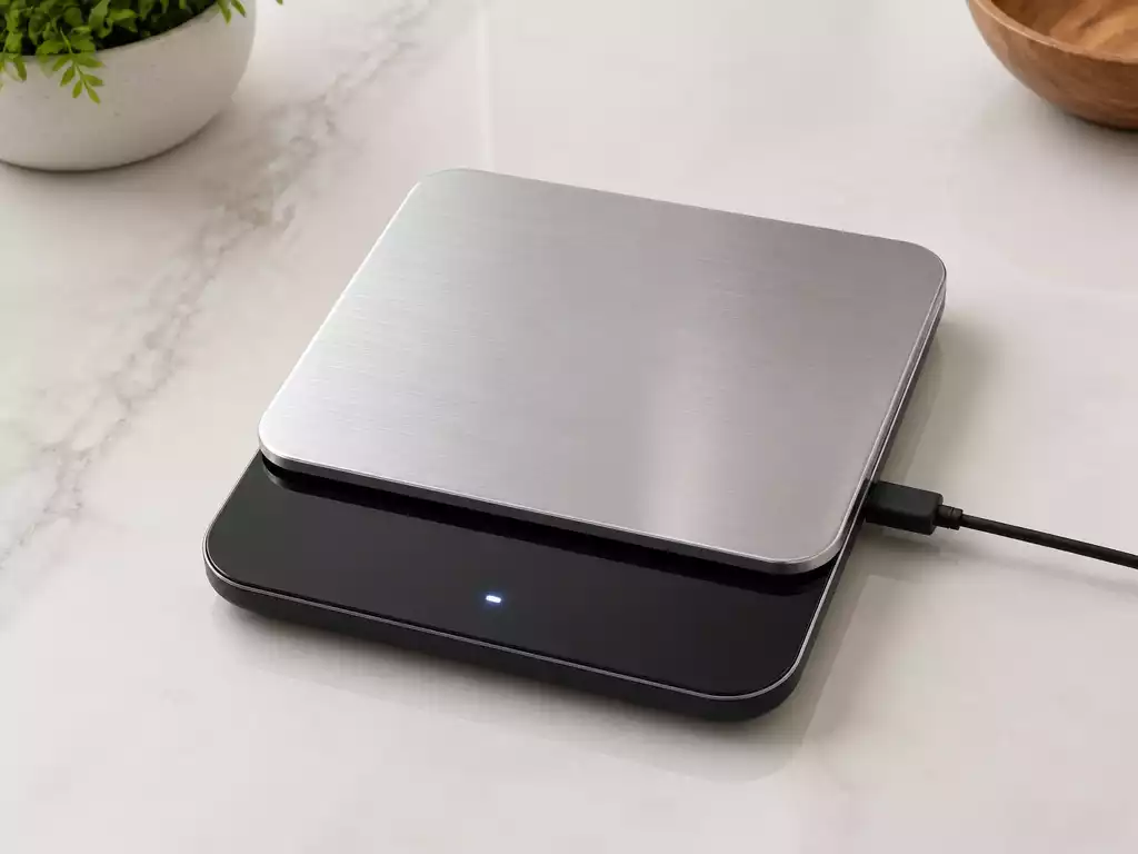 Digital kitchen scale displaying a precise weight, with a charger cable plugged in, on a clean countertop