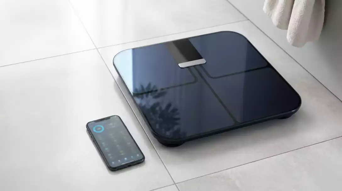 Garmin Index S2 smart scale on a bathroom floor with a smartphone showing the Garmin Connect app data