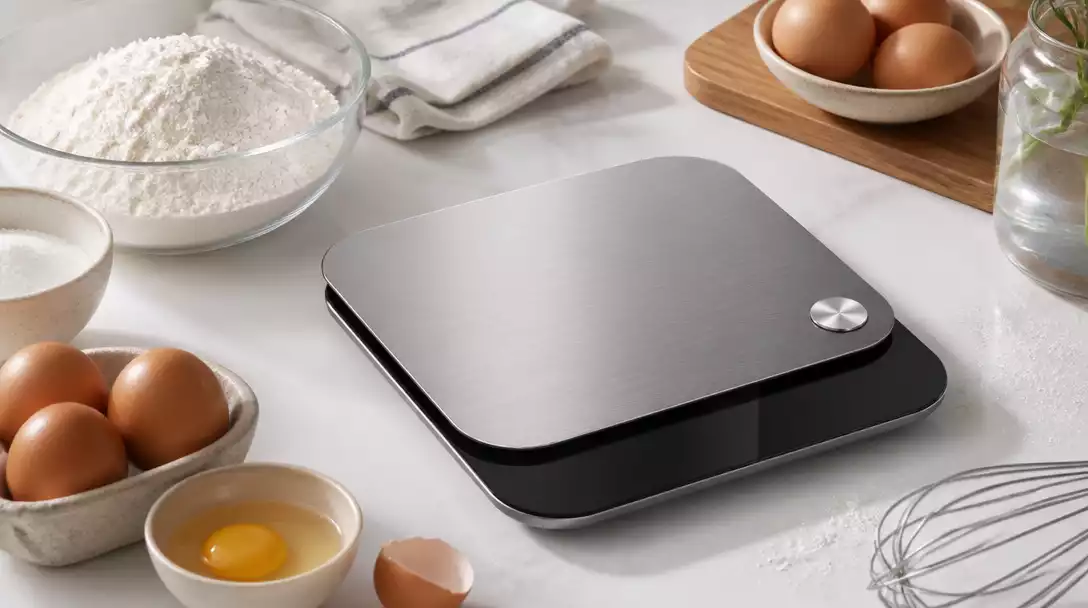 Modern digital kitchen scale on a clean countertop, with fresh ingredients like flour and eggs around it