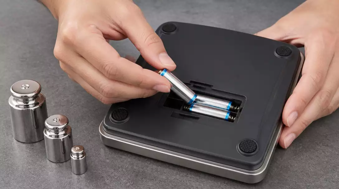 Close-up of hands replacing batteries in a digital scale, with calibration weights nearby