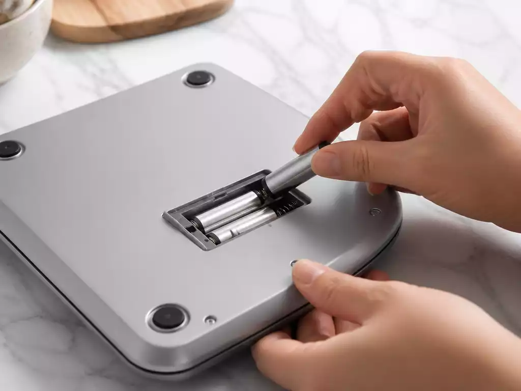 Close-up of hands replacing batteries in a digital kitchen scale on a clean countertop
